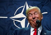Donald Trump speaks at a press conference with a NATO flag in the background.