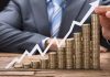 Стовпчики монет, що зростають, та графік зі стрілкою вгору на тлі чоловіка в костюмі.