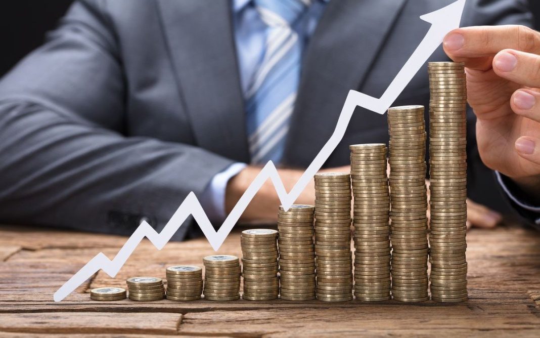 Stacks of coins rising up, and a graph with an upward-pointing arrow, set against the backdrop of a man in a suit.