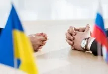 Україна хоче прискорити переговори з РФ та залучити ЄС Представники України та Росії сидять за столом переговорів, на передньому плані розміщені український і російський прапори