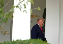 US President Donald Trump, wearing a dark blue suit and a red tie, at the entrance to the White House.