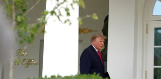US President Donald Trump, wearing a dark blue suit and a red tie, at the entrance to the White House.