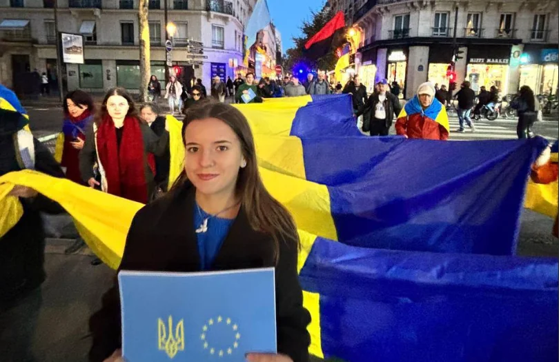 Українські та французькі активісти під час вечірньої ходи в Парижі несуть великі прапори України та плакат із символікою України і Євросоюзу.