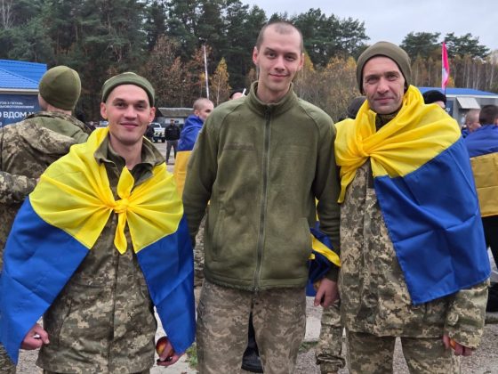 Three Ukrainian servicemen after their release from Russian captivity; two of them are wrapped in blue and yellow flags.