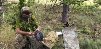 Український військовослужбовець у маскувальному спорядженні налаштовує кабель супутникового термінала Starlink у лісовій місцевості.