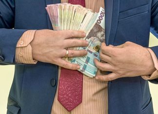 A man in a business suit is holding bundles of Ukrainian hryvnias tucked into his jacket, illustrating the issue of high salaries for officials and budgetary constraints.