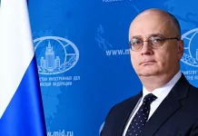Російський дипломат Гліб Десятников у діловому костюмі на тлі символіки Міністерства закордонних справ Росії та державного прапора РФ